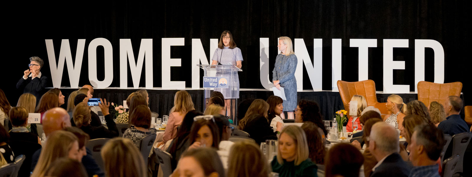 Two women talking in front of a large group at dining tables