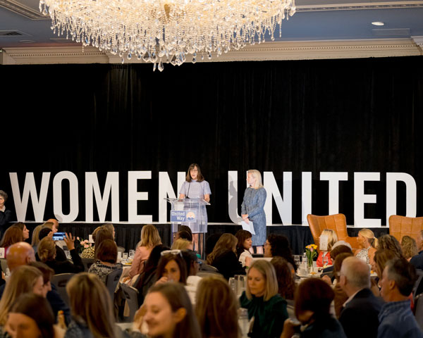 two women on a stage talking to a crowd at tables